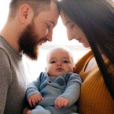a man with a beard and a woman with long dark hair look down at a baby wearing blue between them